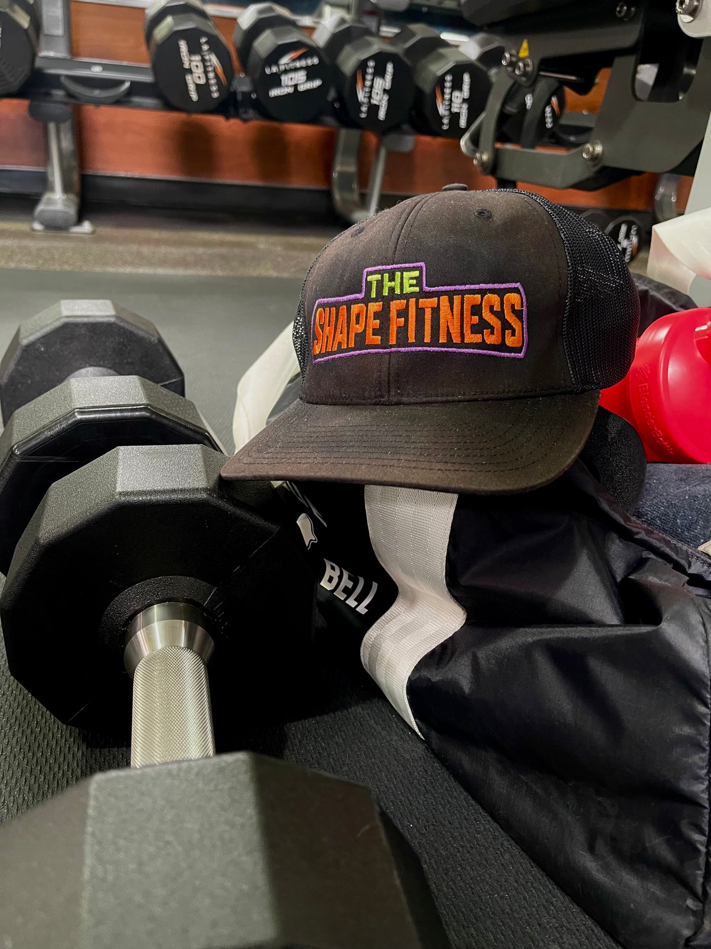 Black cap with 'The Shape Fitness' logo on a gym bag with weights