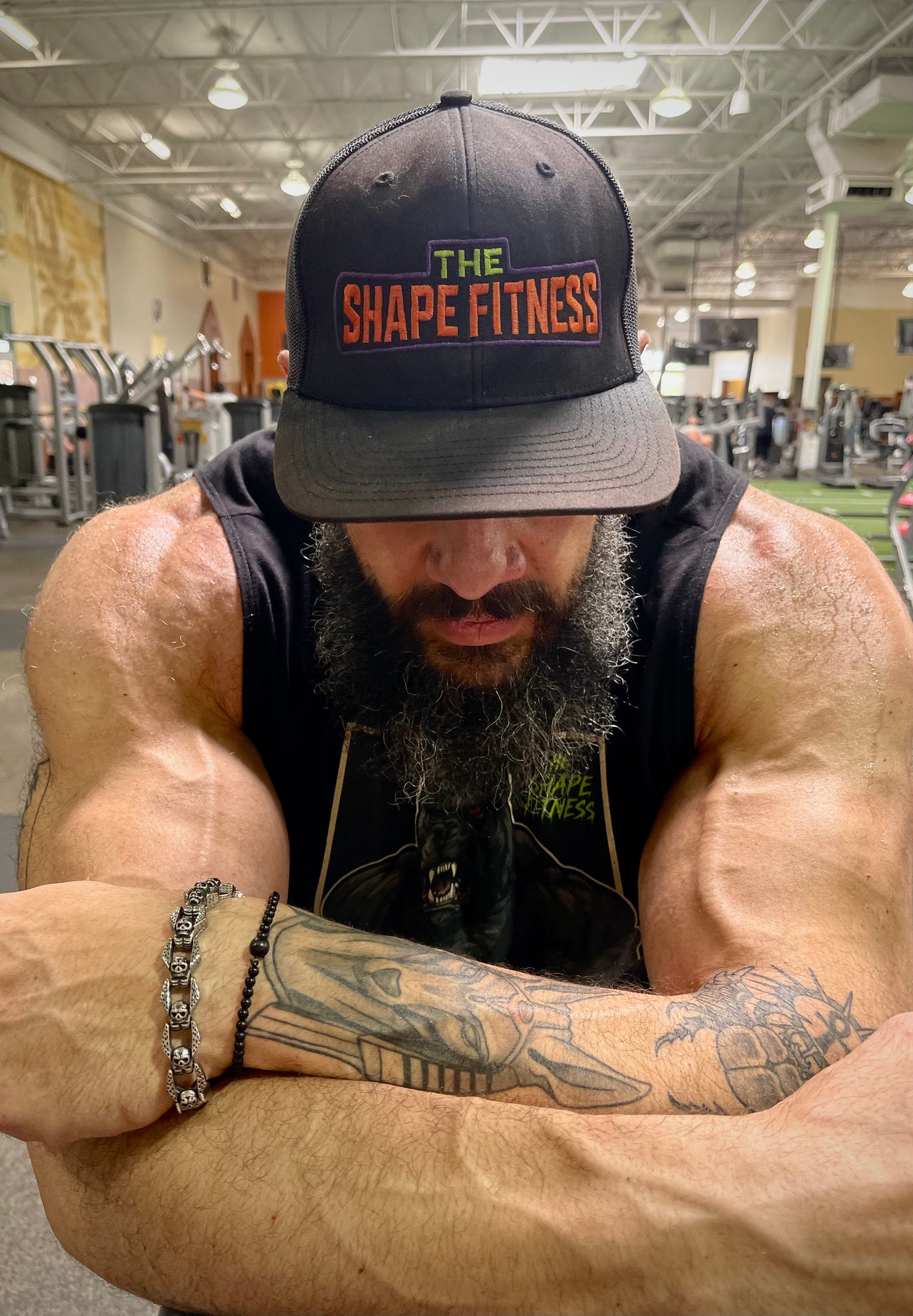 Muscular man in a gym wearing a 'The Shape Fitness' cap.