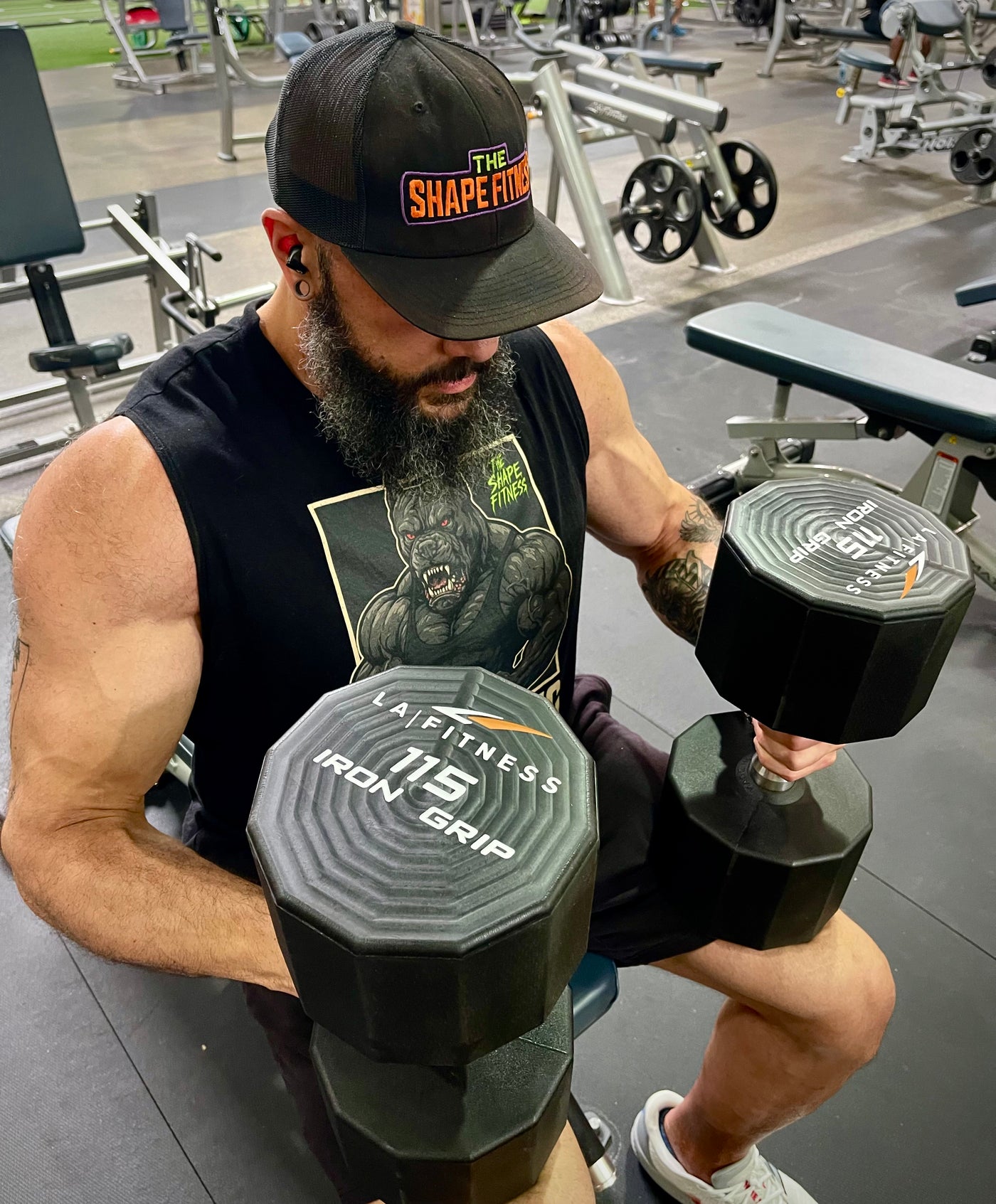 Man in a gym holding dumbbells with 'The Shape Fitness' branded hat